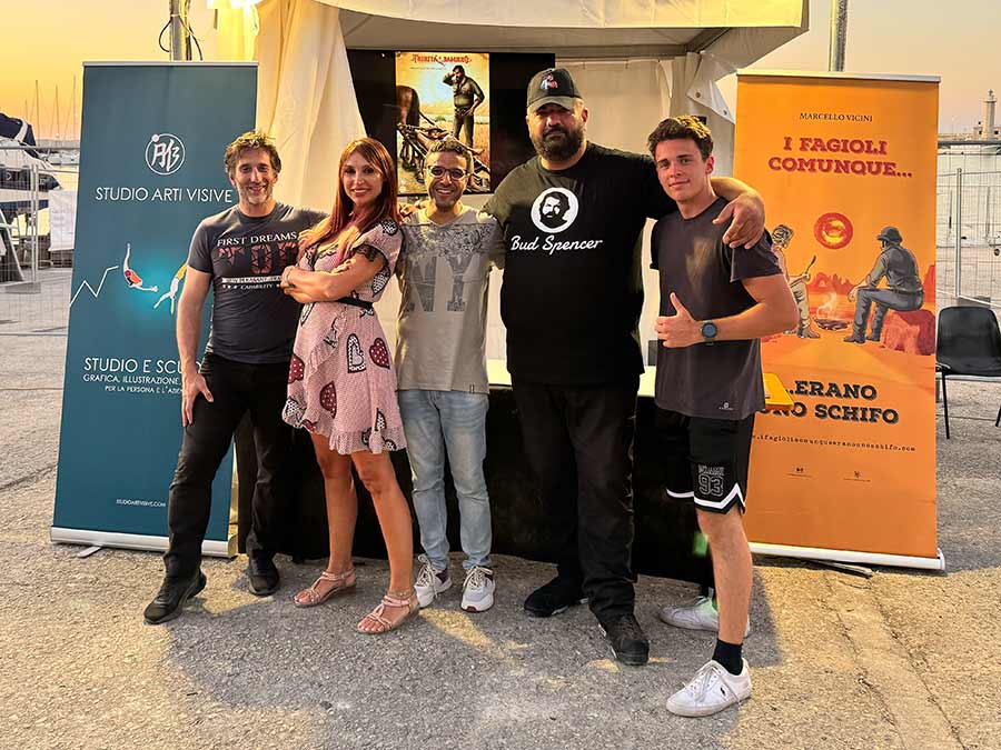 Quattro uomini e una donna posano davanto ad uno stand con ai lati due rollup, sullo sfondo i colori di un tramonto sul mare