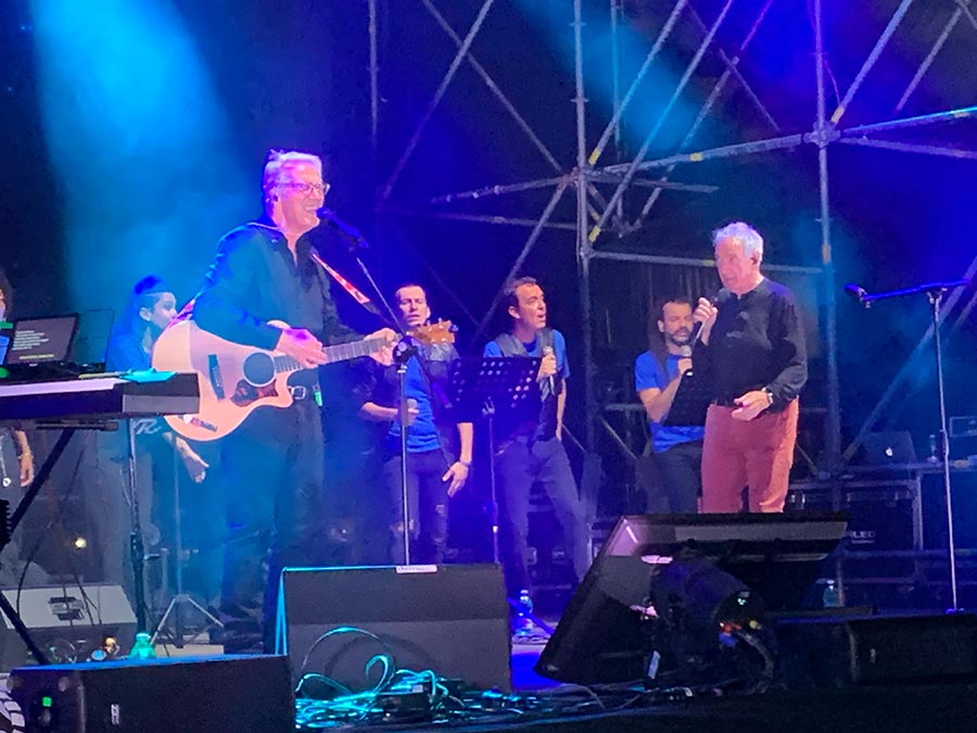 Due cantanti cantano su un palco con alle spalle un coro e davanti casse, microfoni e strumenti musicali