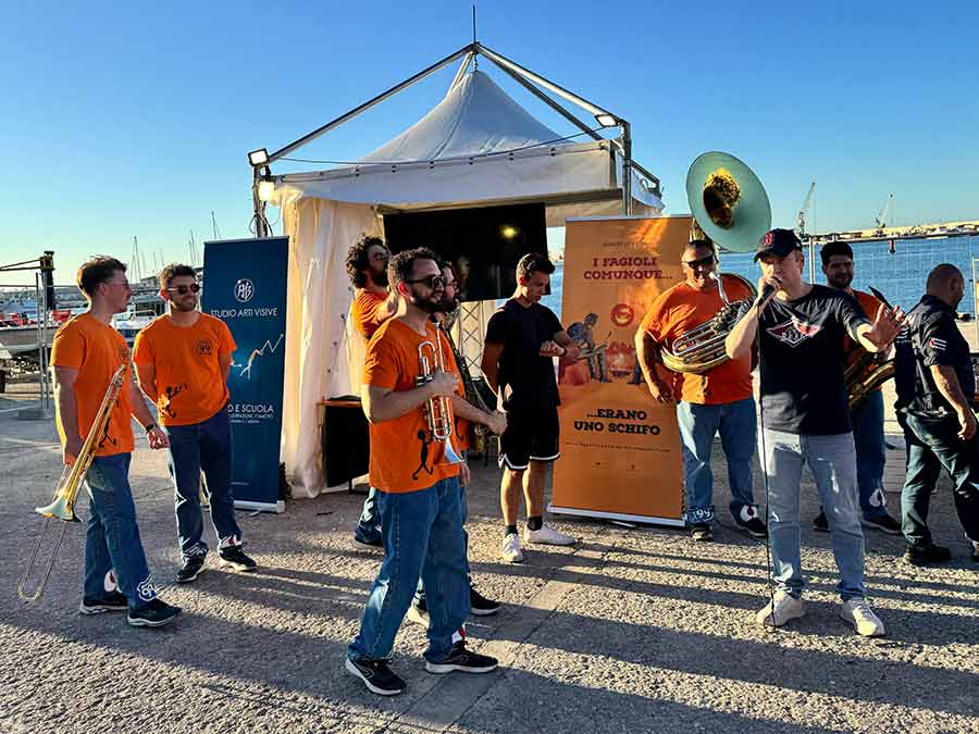 Un uomo al microfono parla con attorno una banda musicale e alle spalle lo sfondo del mare