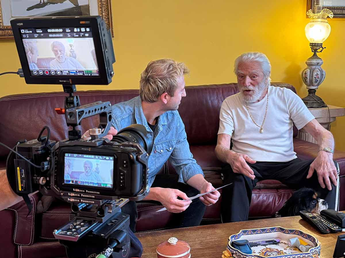 Due uomini seduti su un divano parlano mentre in primo piano una videocamera li riprende