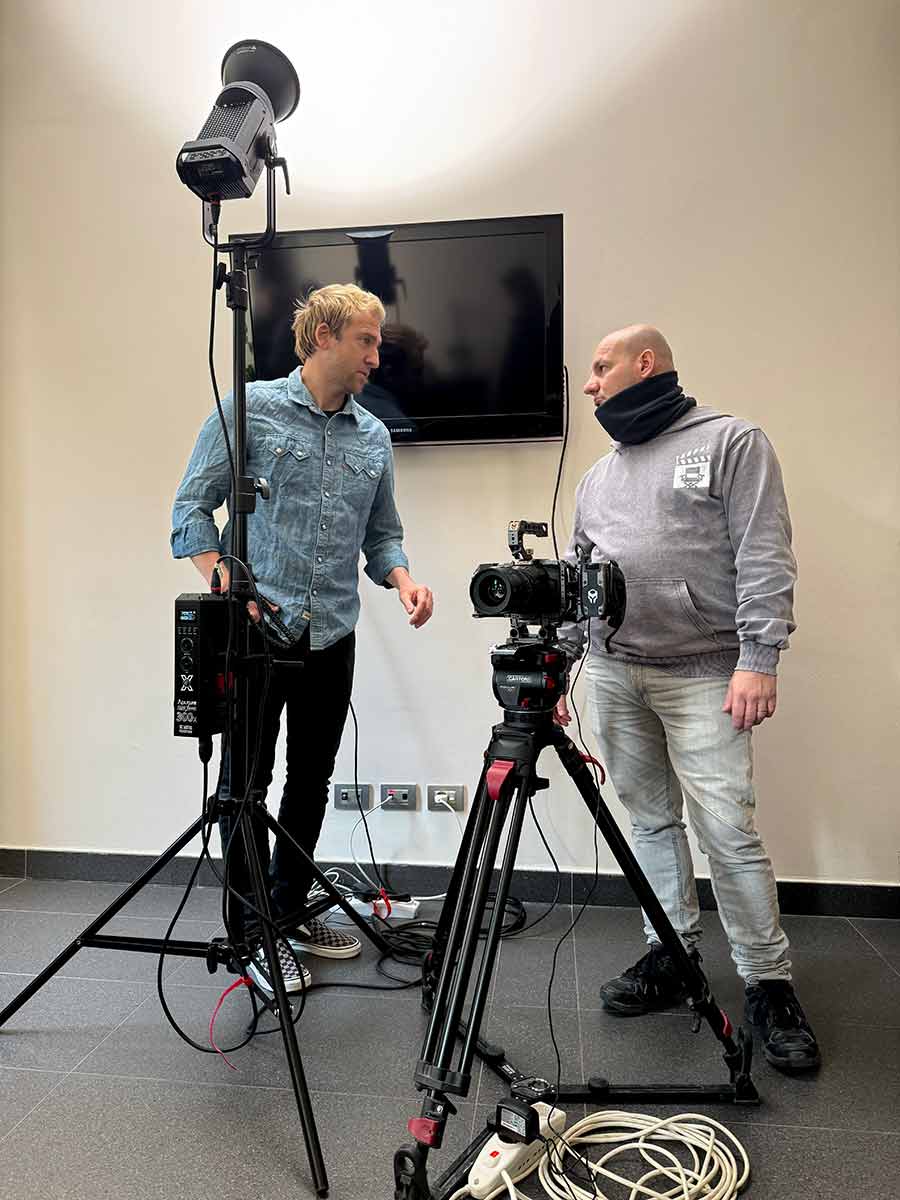 Due uomini in piedi parlano con attorno una attrezzatura in allestimento per un set cinematografico, formata da un faro, una televisione e una videocamera con cavi a terra