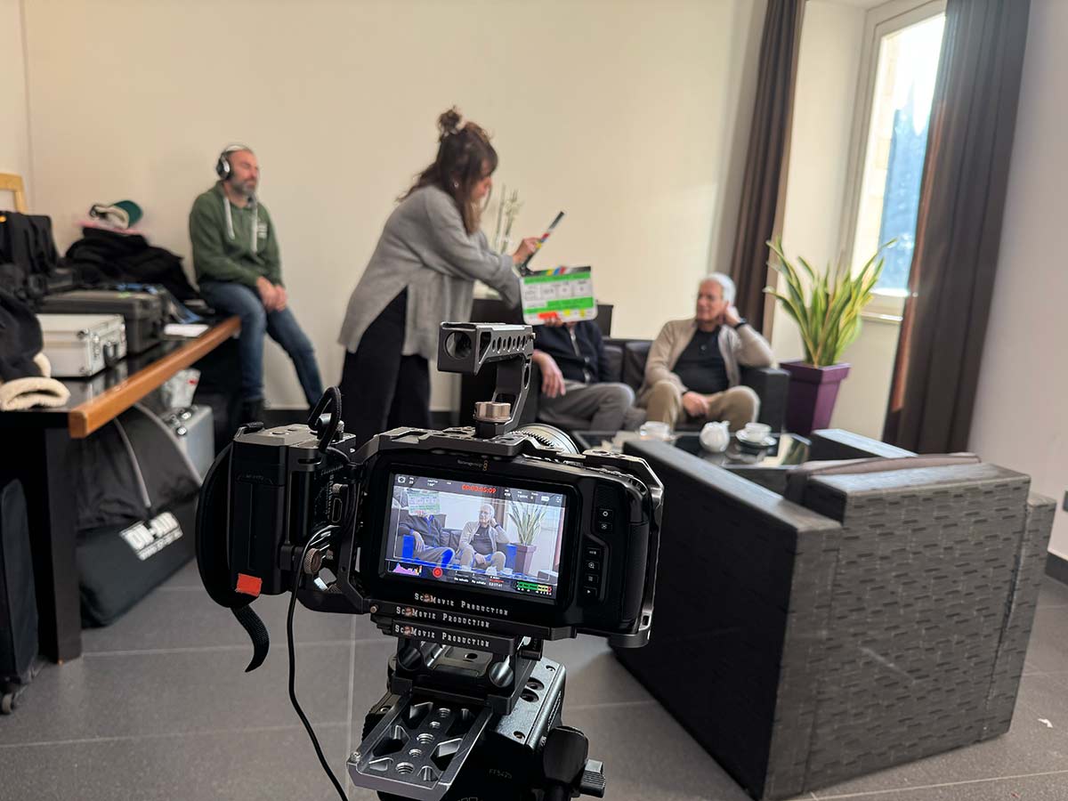 Uno schermo di una videocamera in primo piano riprende sullo sfondo un set con quattro persone che stanno preparando una intervista all'interno di un salottino